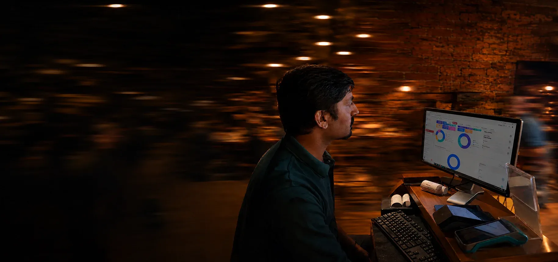 Restaurant cashier at a POS station reviewing the Restaurant OS dashboard on a monitor